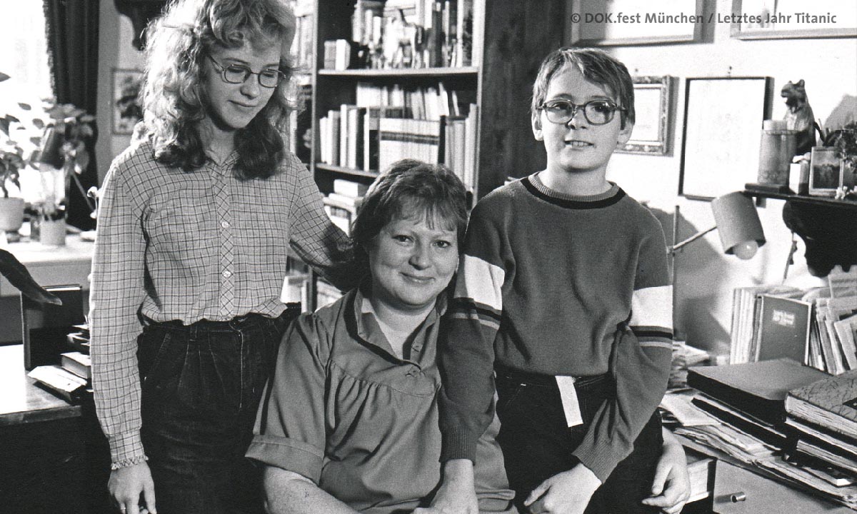 Filmstill aus "Letztes Jahr Titanic": Schwarz-weiß-Bild einer Mutter mit zwei Kindern in einem Zimmer voller Bücher (© DOK.Fest München / Letztes Jahr Titanic)