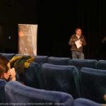 Bild mehrerer Personen mit Mikrofon vor einer Leinwand