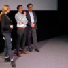 Bild von drei Personen mit Mikrofon vor einer Leinwand