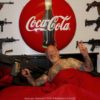 Oberkörperfreier Mann im Bett, umgeben von mehreren Maschinengewehre mit Coca Cola Logo im Hintergrund