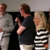 Bild mehrerer Personen mit Mikrofon vor der Leinwand