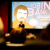 Foto von der Kino-Premiere von ROBIN BANK in Stuttgart. Zu sehen ist Arek Gielnik (Indi Film), der vor der Leinwand steht und das Kinopublikum begrüßt. Die Menschen im Kino-Saal sind unscharf abgebildet, die Kamera fokussiert von hinten Richtung Leinwand. © Indi Film