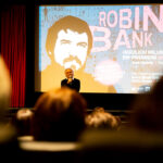 Foto von der Kino-Premiere von ROBIN BANK in Stuttgart. Zu sehen ist Arek Gielnik (Indi Film), der vor der Leinwand steht und das Kinopublikum begrüßt. Die Menschen im Kino-Saal sind unscharf abgebildet, die Kamera fokussiert von hinten Richtung Leinwand. © Indi Film