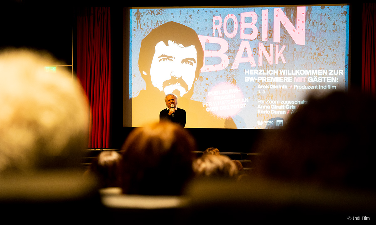 Foto von der Kino-Premiere von ROBIN BANK in Stuttgart. Zu sehen ist Arek Gielnik (Indi Film), der vor der Leinwand steht und das Kinopublikum begrüßt. Die Menschen im Kino-Saal sind unscharf abgebildet, die Kamera fokussiert von hinten Richtung Leinwand. © Indi Film