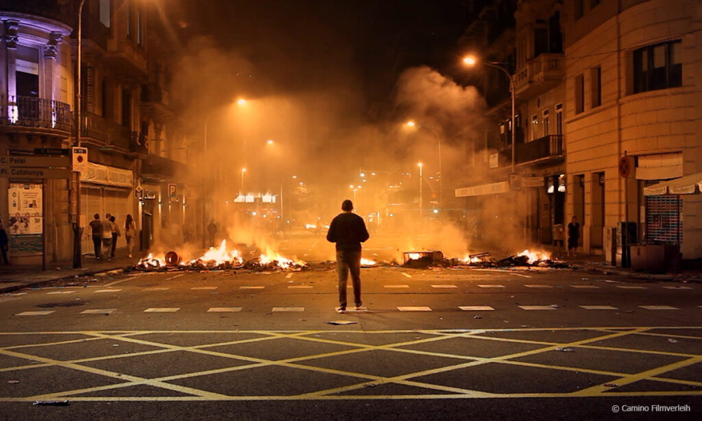Filmstill aus ROBIN BANK. Abgebildet ist eine Person von hinten, die auf brennende Straßenbarrikaden während Unruhen schaut. © Camino Filmverleih