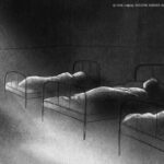 Filmstill aus DIE KINDER AUS KORNTAL. Man sieht ein animiertes Bild in Schwarz-Weiß, das einen Schlafsaal mit drei Betten zeigt sowie eine stehende Person am Rand des Bildes, die von hinten dargestellt wird und etwas betrachtet oder rausschaut. Eine genaue Einordnung des Blickes ist schwer möglich. (Credit: DOK Leipzig 2023/Regie: Julia Charakter)