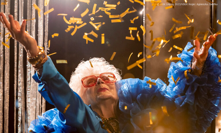 Filmstill aus VIKA! Man sieht eine ältere Frau im Partyoutfit mit großer roter Brille, die goldenes Konfetti wirft und offensichtlich eine gute Zeit hat (Credit: DOK LEIPZIG 2023/Regie: Agnieska Zwiefa)